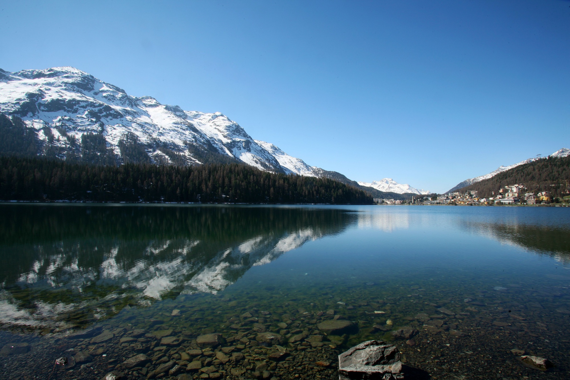 View of snow-capped mountains reflected in a lake under a clear blue sky in St. Moritz, Switzerland.